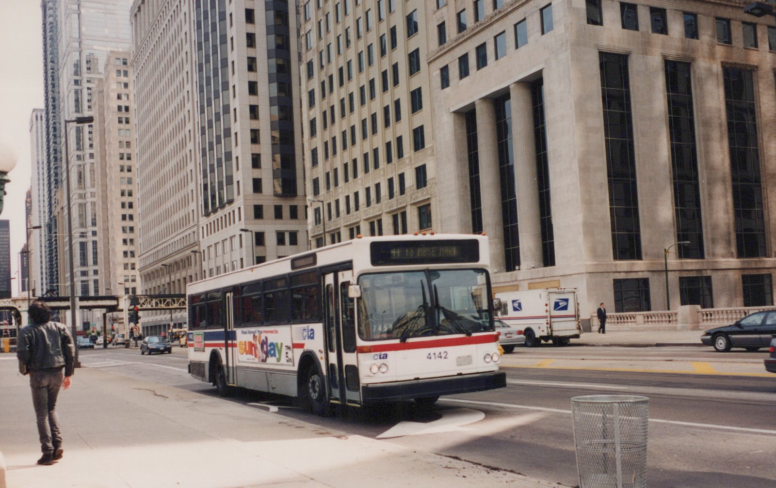 A white and red CTA bus with the number 4142 on the front is driving on a city street with tall buildings in the background and a pedestrian on the sidewalk.