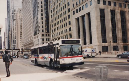 A white and red CTA bus with the number 4142 on the front is driving on a city street with tall buildings in the background and a pedestrian on the sidewalk.