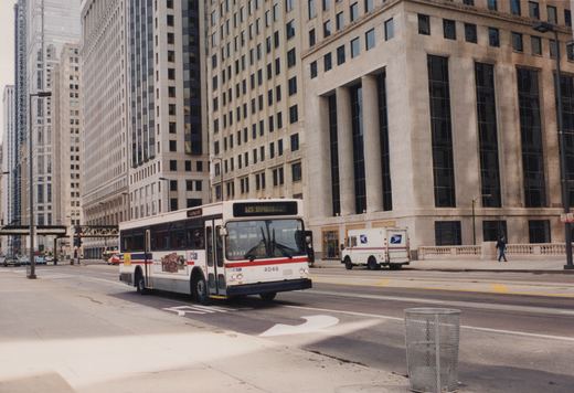 A white CTA bus with red and blue stripes, numbered 4046, is driving down a wide city street lined with tall office buildings, with a postal truck parked on the sidewalk nearby.
