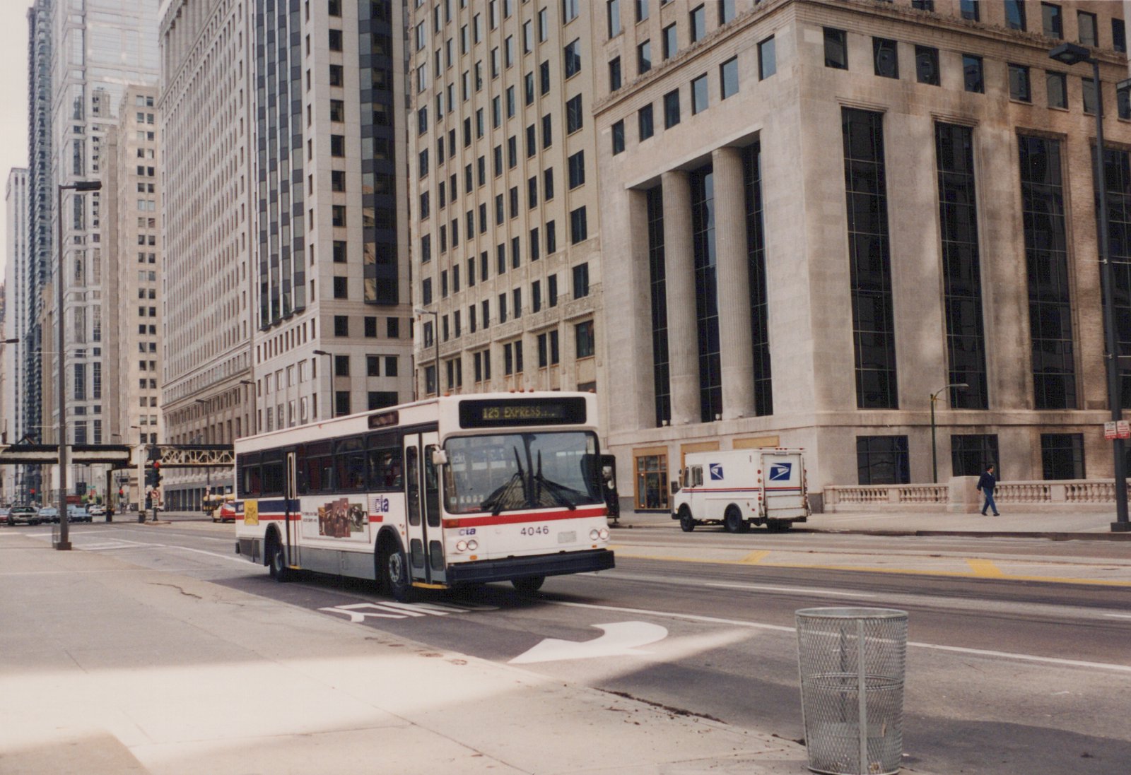 A white CTA bus with red and blue stripes, numbered 4046, is driving down a wide city street lined with tall office buildings, with a postal truck parked on the sidewalk nearby.