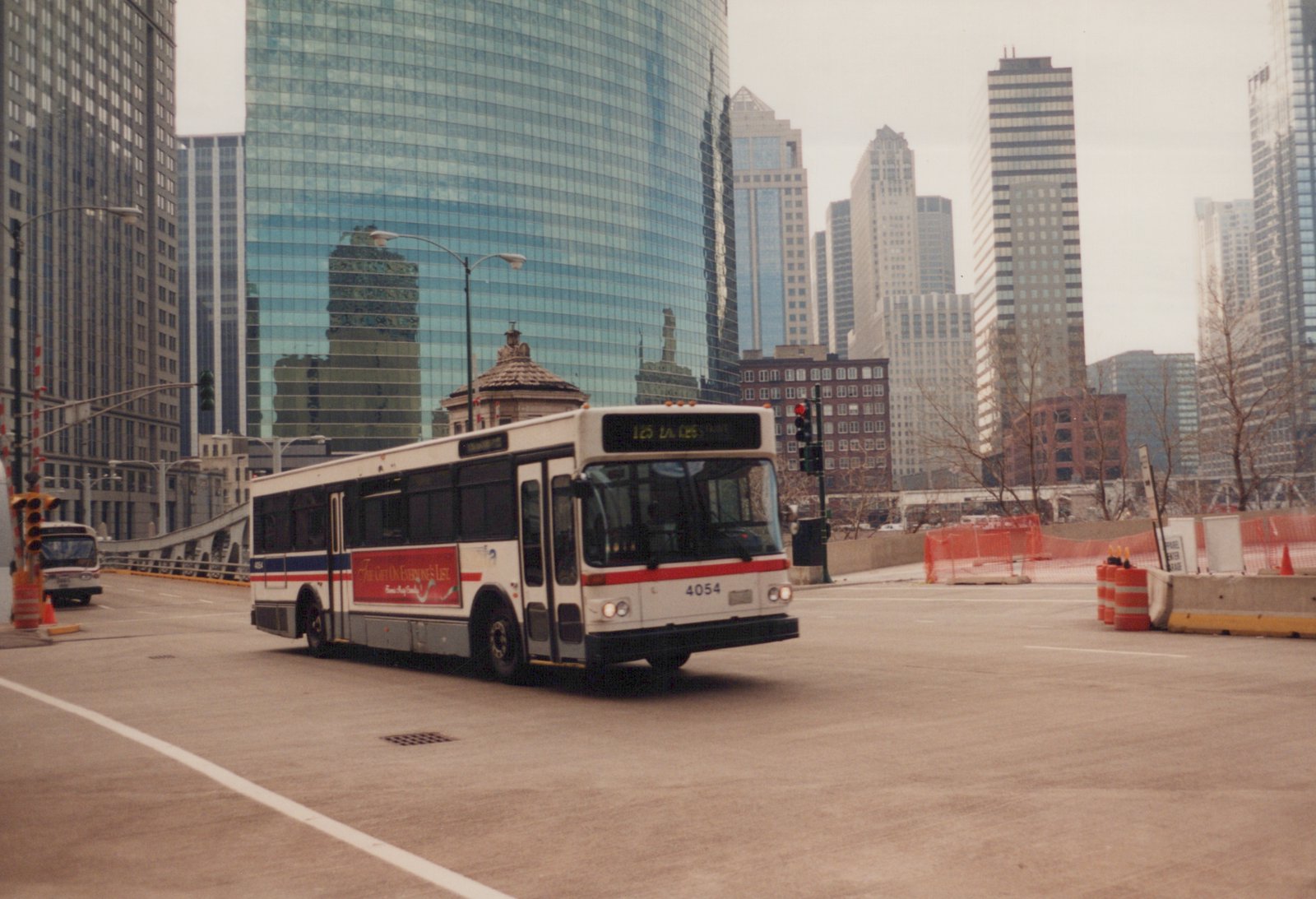 A white Chicago Transit Authority bus with the number 4054 and destination 125 LAKE is driving on a city street with tall buildings in the background.