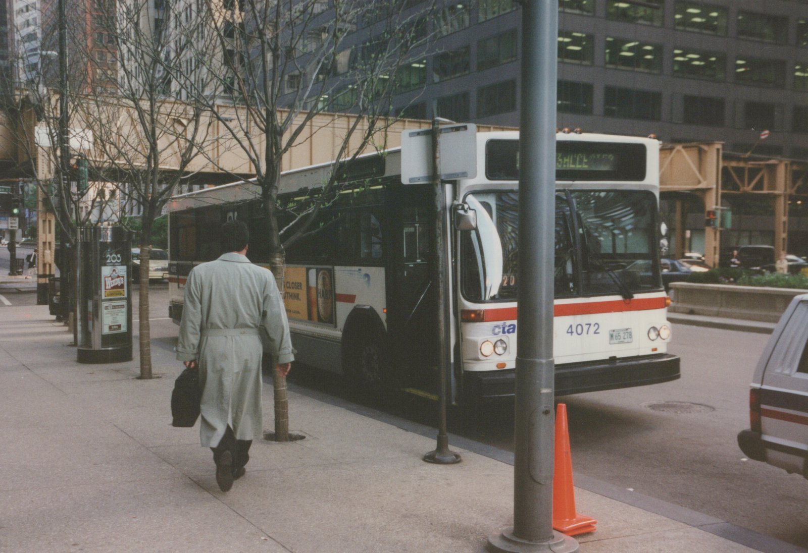 Front three-quarter view of a white CTA bus with orange stripe, number 4072, at a city street stop. A man in a long coat walks on the sidewalk, and an elevated railway is in the background.