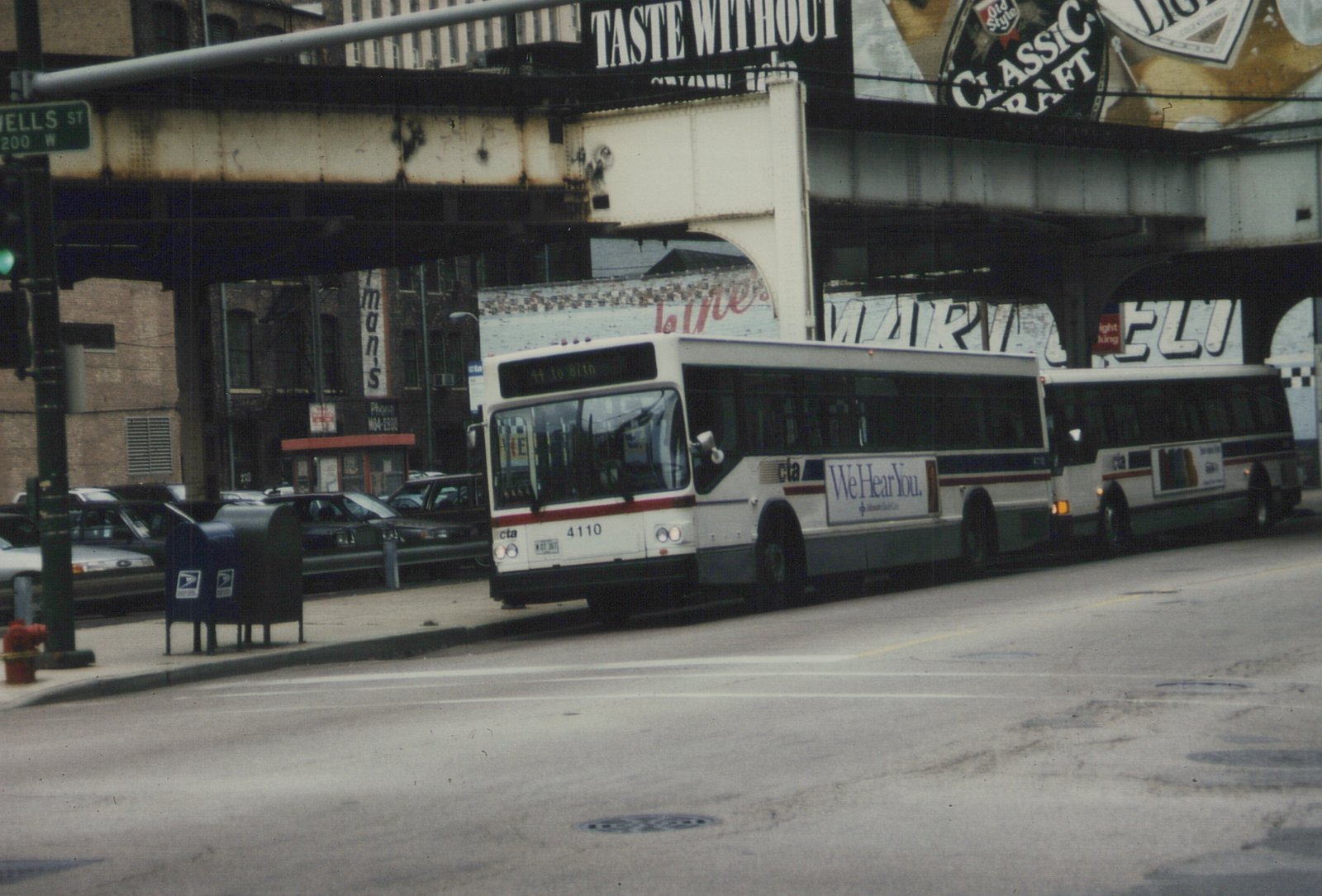 Two white and blue Chicago Transit Authority buses drive down a city street under an elevated train track, with a street sign for Wells Street visible on the left.