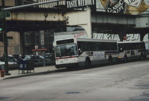 Two white and blue Chicago Transit Authority buses drive down a city street under an elevated train track, with a street sign for Wells Street visible on the left.
