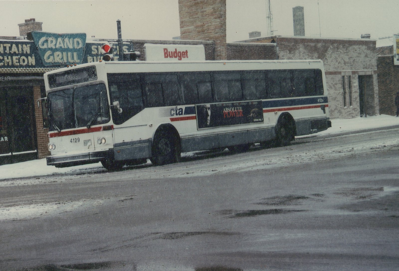 A white and blue CTA bus with the number 4129 is parked on a snow-covered street, with a 'Grand Grill' sign and a 'Budget' sign visible on the building behind it.
