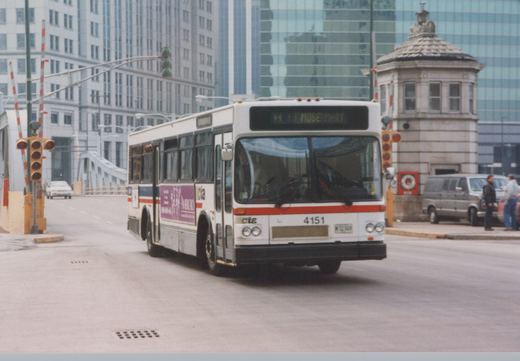 A white Chicago Transit Authority bus with red and blue stripes, numbered 4151, is driving on a city street near a bridge with modern buildings in the background. A grade crossing signal is visible on the left, and two people stand on the sidewalk to the right.