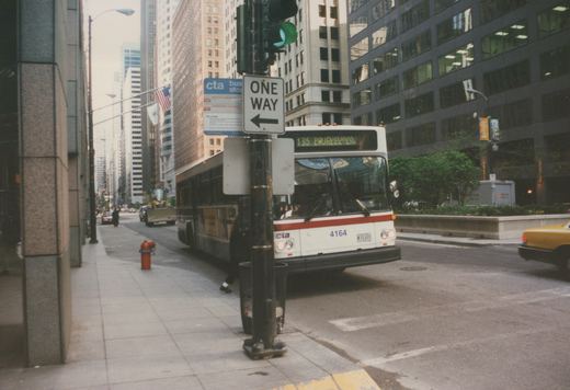 A white CTA bus with the number 4164 is stopped at a bus stop on a city street in Chicago, with a '135 LA SALLE' destination sign, surrounded by tall buildings and a one-way sign.