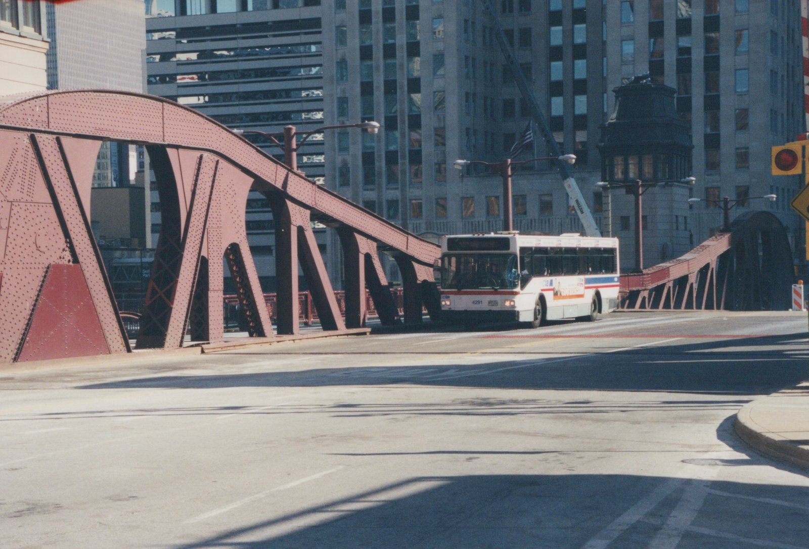 A white and blue Chicago Transit Authority bus, number 4291, is driving on a city street over a red truss bridge, with tall buildings in the background.