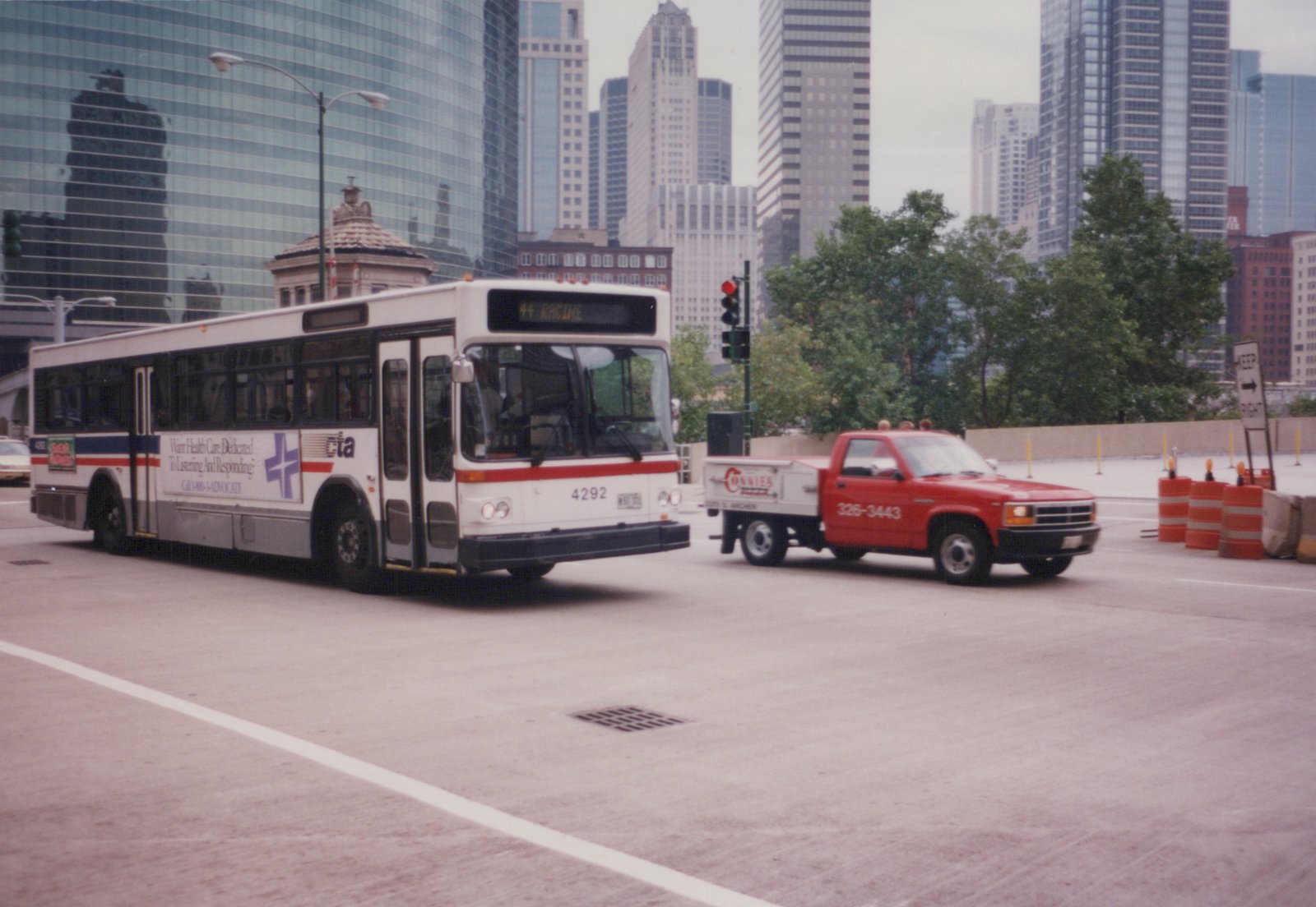 A white Chicago Transit Authority bus with red and blue stripes, number 4292, is driving on a city street with tall buildings behind it, while a red pickup truck is visible nearby.