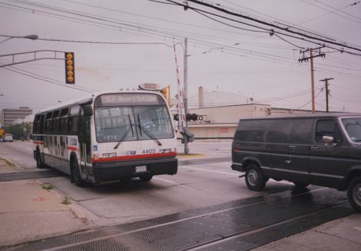 A white CTA bus with red and blue stripes, number 4405, is stopped at a railroad crossing on a wet, overcast day, with a black van crossing in front of it.