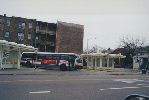 A white, blue, and red Chicago Transit Authority bus numbered 4464 is stopped at a covered bus shelter on a street, with a brick building in the background under an overcast sky.
