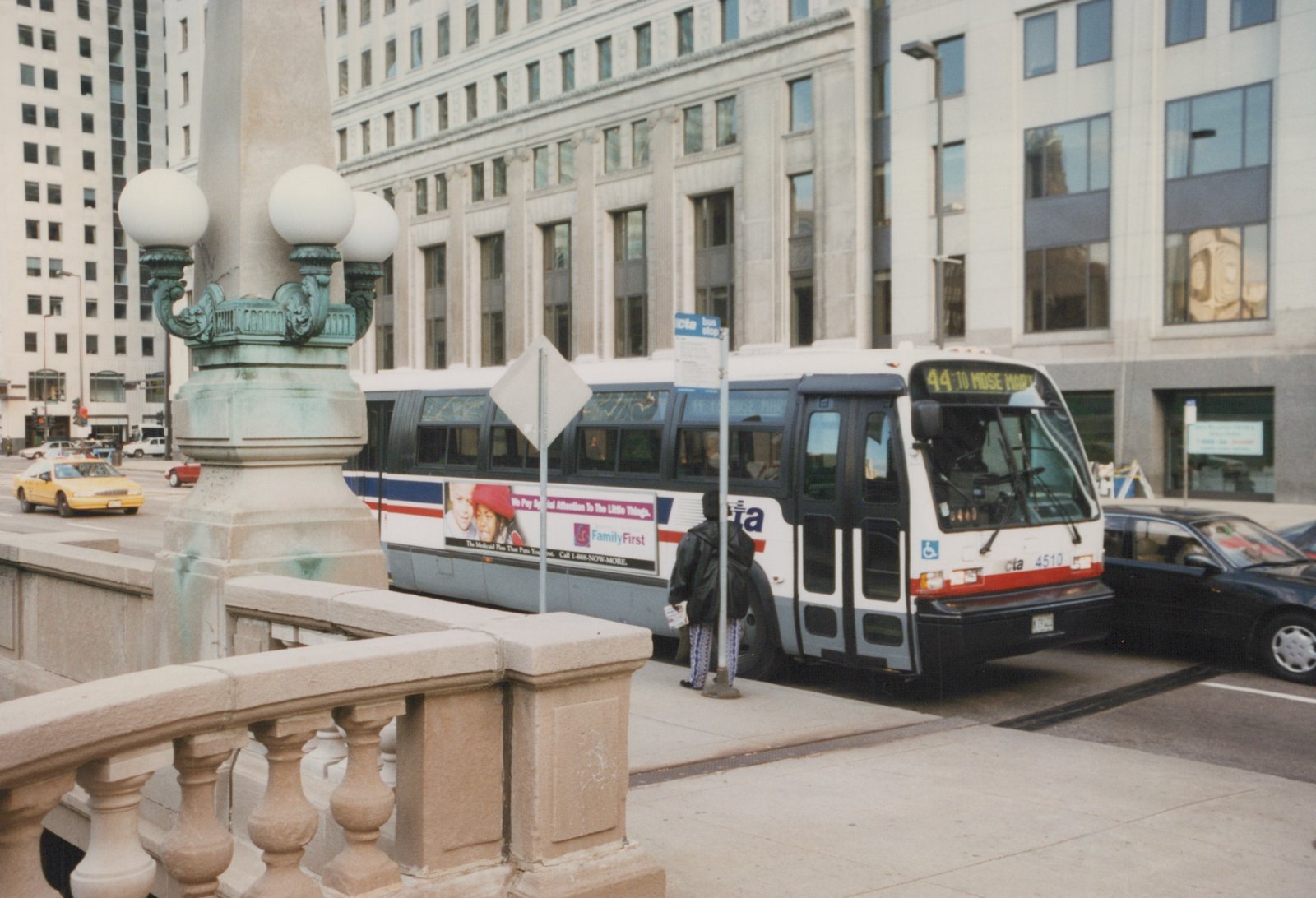 A white, red, and blue CTA bus with the number 4510 on the front is stopped at a bus stop on a city street. A person is standing nearby, and there are buildings and cars in the background.