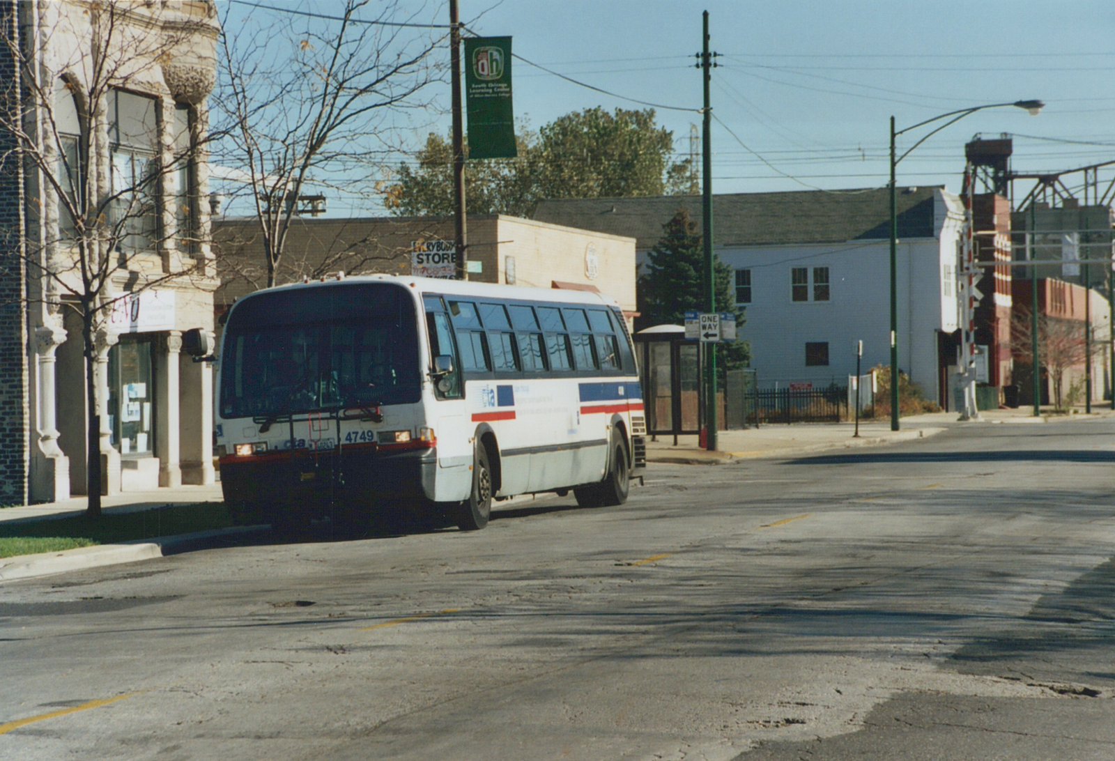 A white CTA bus with blue and red stripes, numbered 4749, drives down a sunlit city street lined with buildings and a bus shelter.