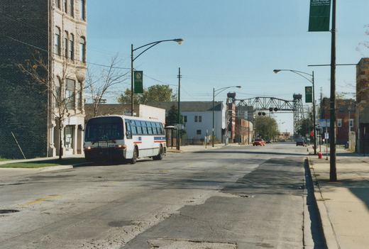A white Pace bus with red and blue stripes is parked on a street corner in South Chicago, near a green banner for the South Chicago Learning Center, with a clear blue sky above.