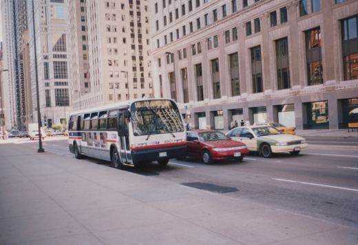 A white Chicago Transit Authority bus with red and blue stripes is driving down a city street past a red car and a white and green taxi, with tall buildings in the background.