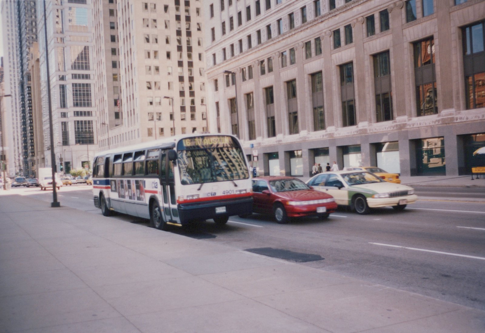 A white Chicago Transit Authority bus with red and blue stripes is driving down a city street past a red car and a white and green taxi, with tall buildings in the background.