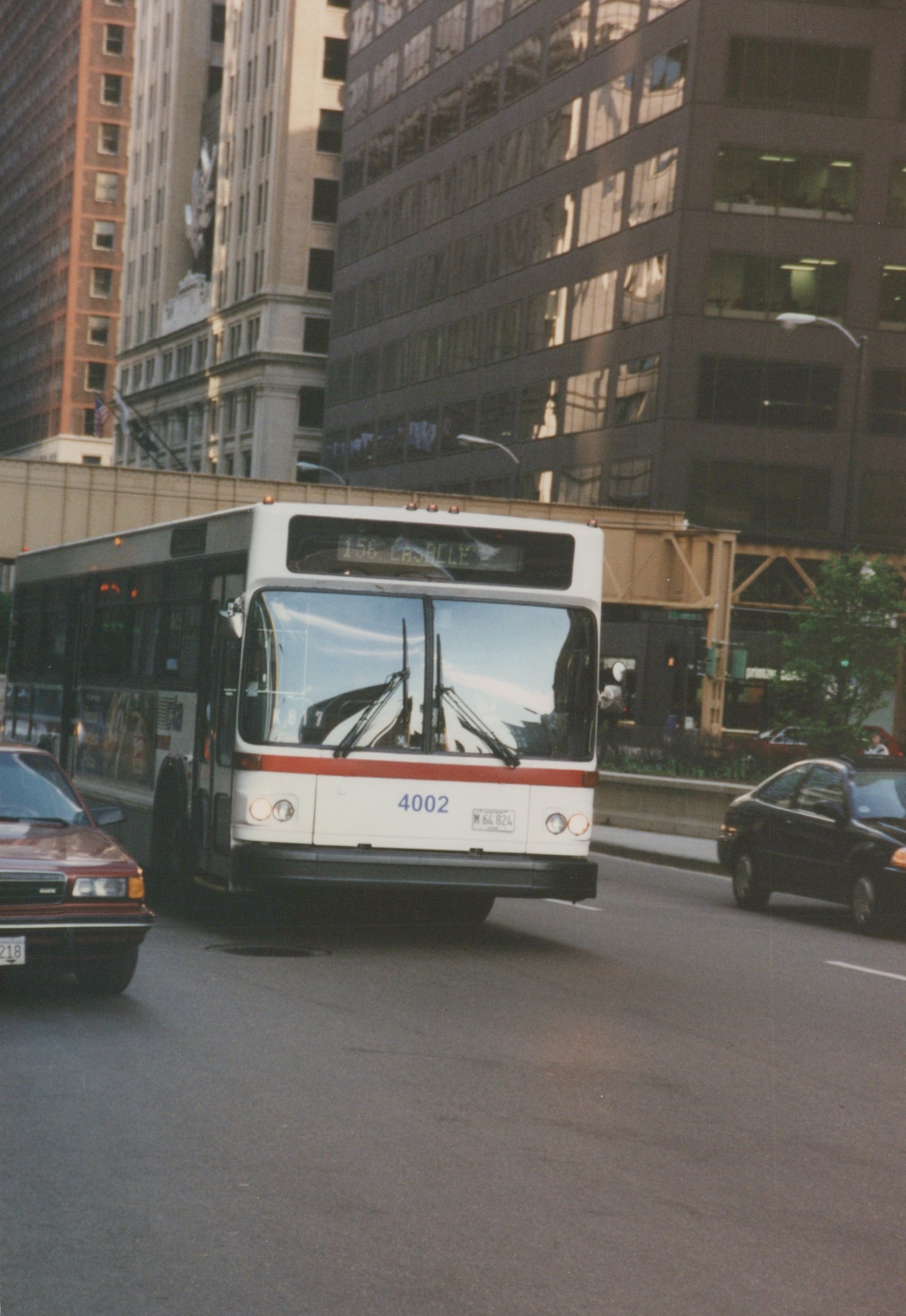 A white and red city bus with the number 4002 is driving down a street in a city, with tall buildings and an elevated rail structure in the background.