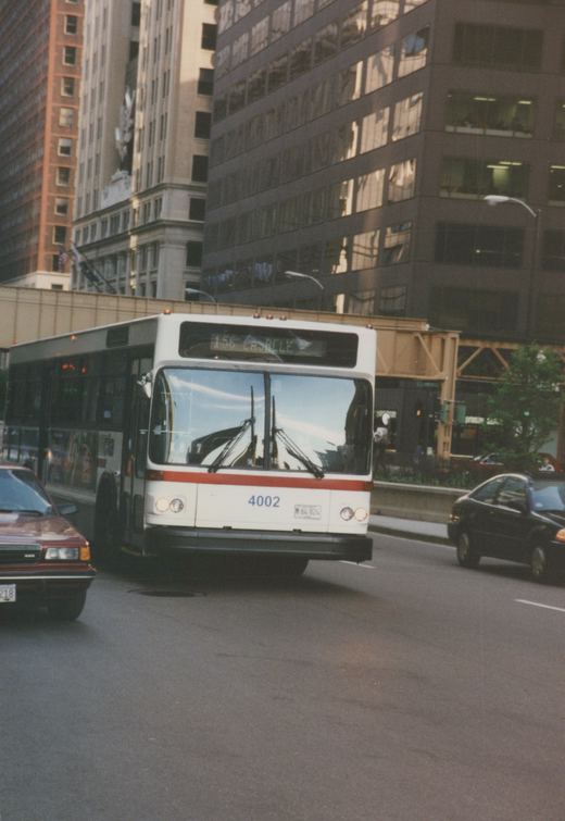 A white and red city bus with the number 4002 is driving down a street in a city, with tall buildings and an elevated rail structure in the background.