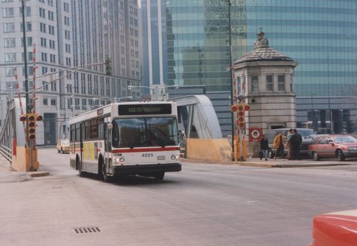 A white CTA bus with red stripe, number 4025, is driving down a city street toward a bridge, with several people and other vehicles nearby.