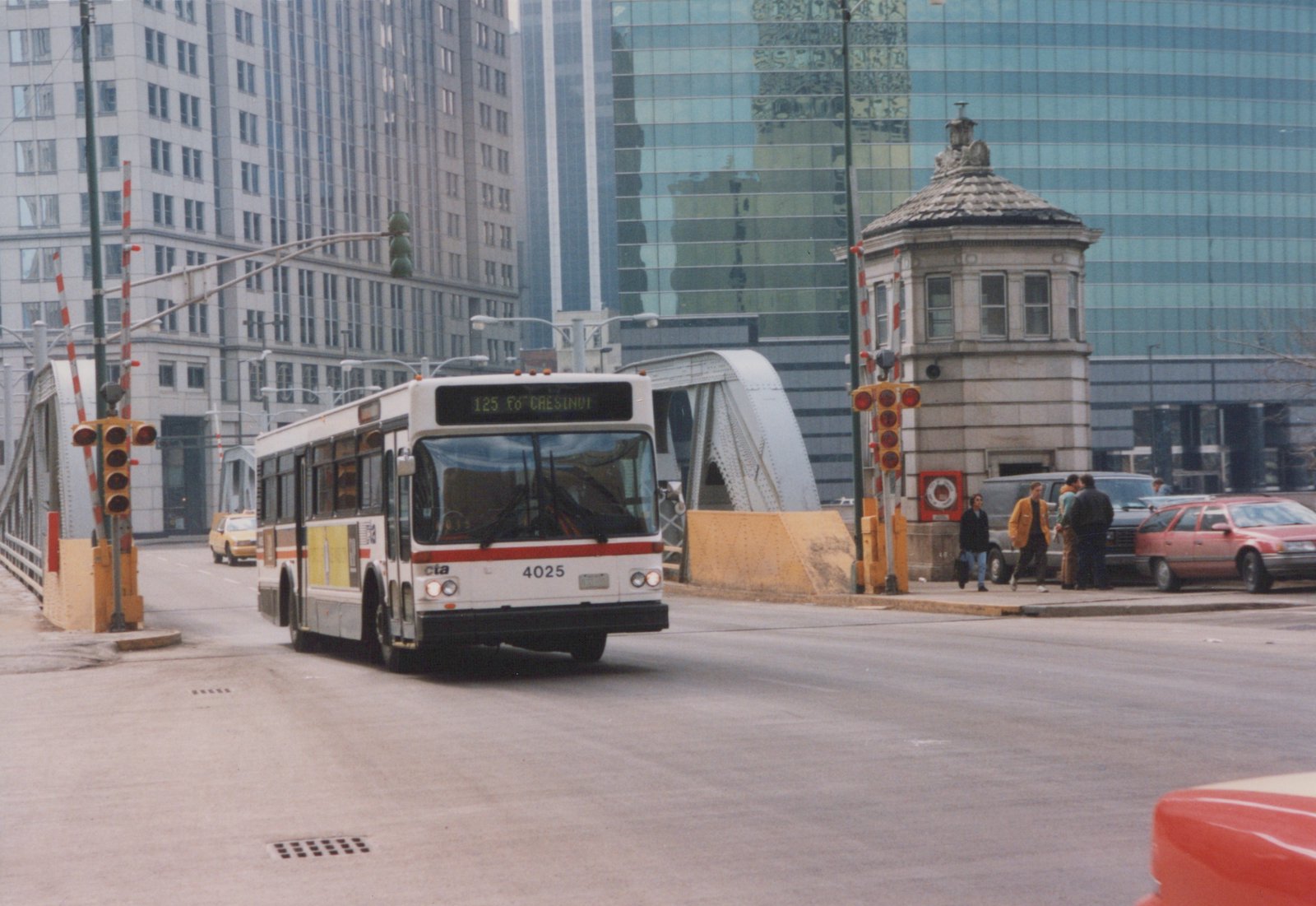 A white CTA bus with red stripe, number 4025, is driving down a city street toward a bridge, with several people and other vehicles nearby.