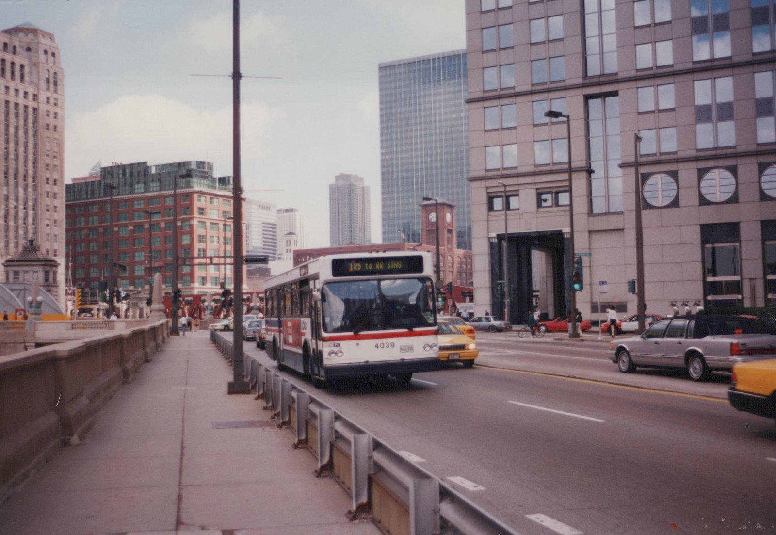A white and red Chicago Transit Authority bus, number 4039, is driving on a multi-lane bridge in the city, with modern buildings and traffic in the background.