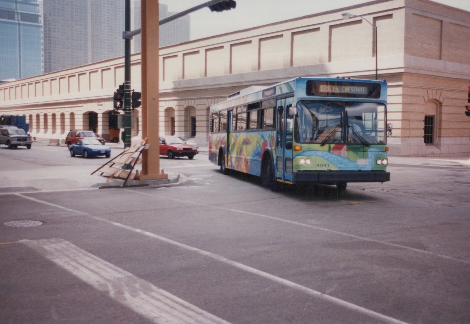 A blue and green city bus with a colorful side mural, numbered 4041, is turning at a street intersection with modern buildings and other cars in the background.