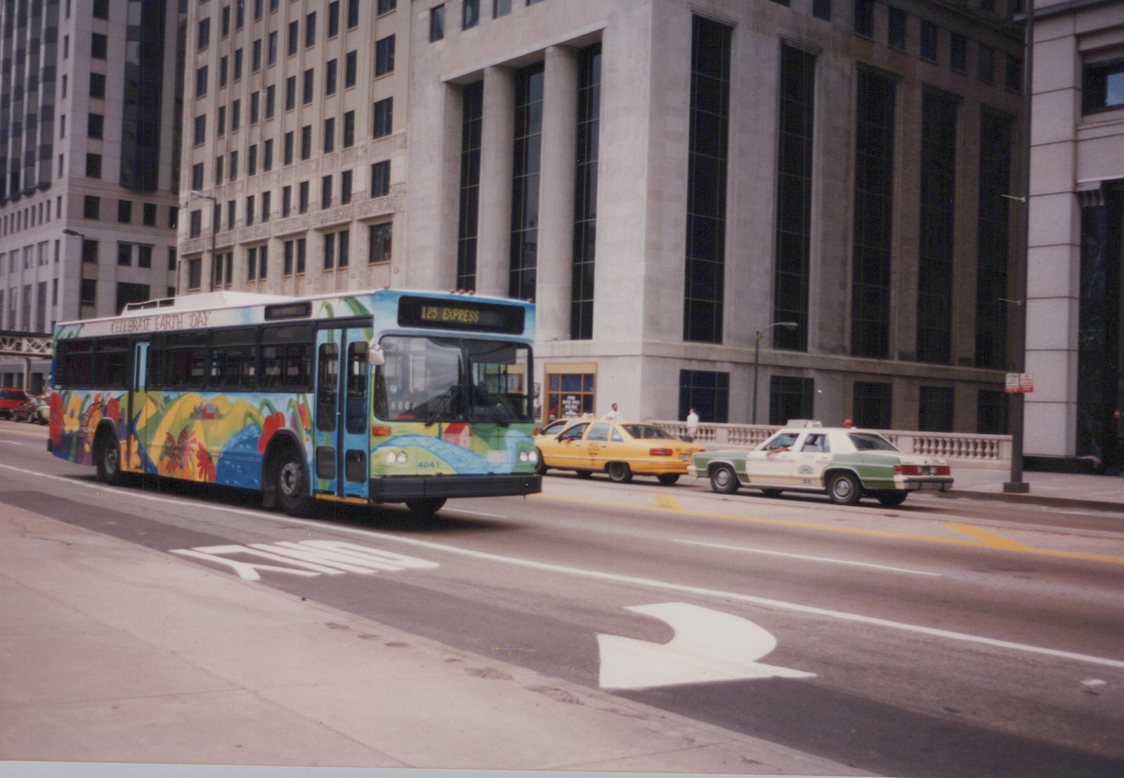 A blue, green, and yellow Chicago Transit Authority bus with an Earth Day design, displaying route 125 Express, drives down a street with taxis and tall buildings in the background.