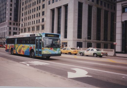A blue, green, and yellow Chicago Transit Authority bus with an Earth Day design, displaying route 125 Express, drives down a street with taxis and tall buildings in the background.