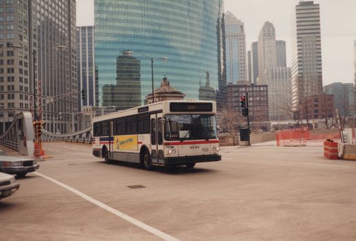 A white, red, and blue Chicago Transit Authority bus with the number 4044 is turning on a city street, set against a backdrop of tall glass and steel skyscrapers on an overcast day.