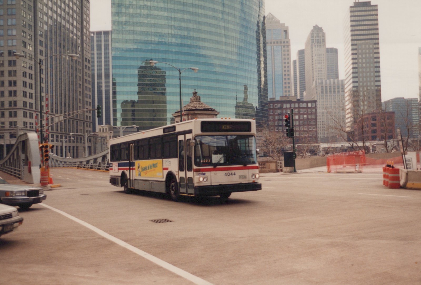 A white, red, and blue Chicago Transit Authority bus with the number 4044 is turning on a city street, set against a backdrop of tall glass and steel skyscrapers on an overcast day.
