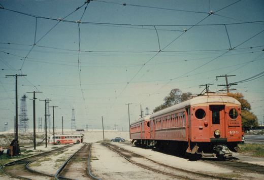 Two red interurban cars with round end windows and trolley poles stand on a yard track under a web of overhead wires, with a red and white bus, a parked car and oil derricks in the distance.