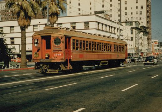 A red interurban car with round front windows runs on street track past a palm tree and tall white buildings, with 1950s cars in the traffic behind it.
