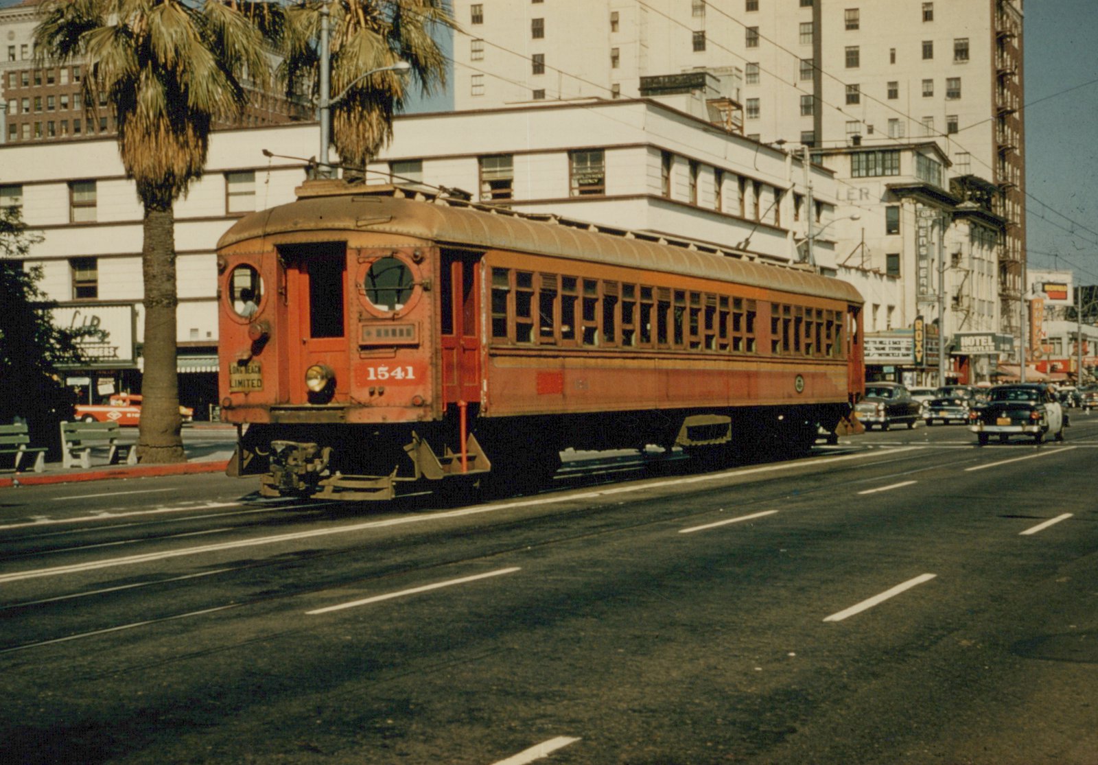 A red interurban car with round front windows runs on street track past a palm tree and tall white buildings, with 1950s cars in the traffic behind it.
