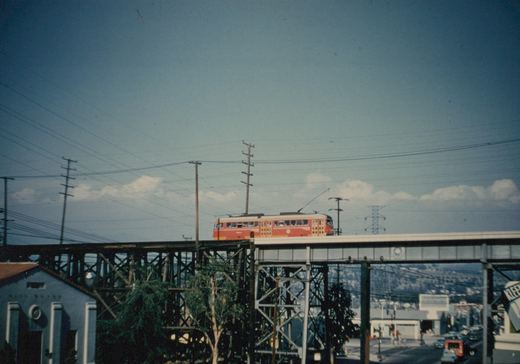 A red and yellow electric car with a trolley pole crosses a long wooden and steel trestle high above a street, with power lines and a blue sky with clouds above.