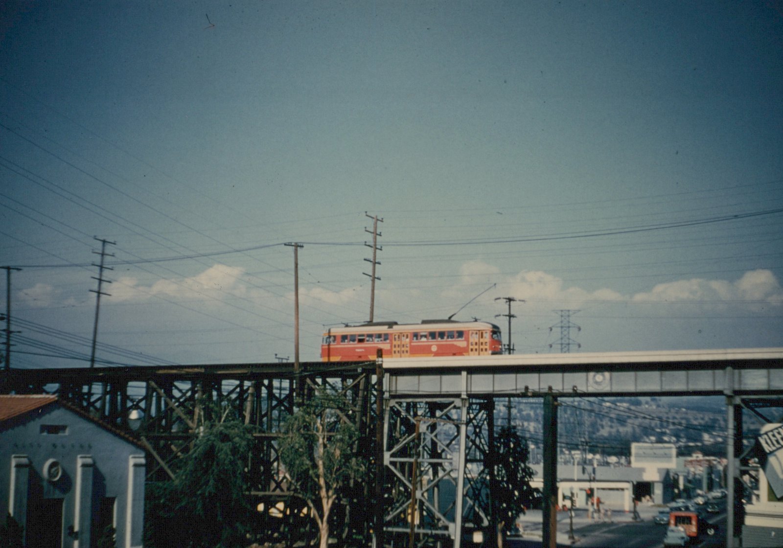 A red and yellow electric car with a trolley pole crosses a long wooden and steel trestle high above a street, with power lines and a blue sky with clouds above.