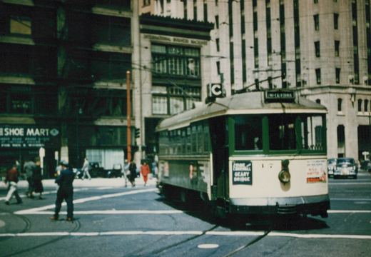 A cream and green streetcar with a trolley pole and a C route sign turns across a downtown intersection with tracks, while pedestrians cross in front of tall office buildings.