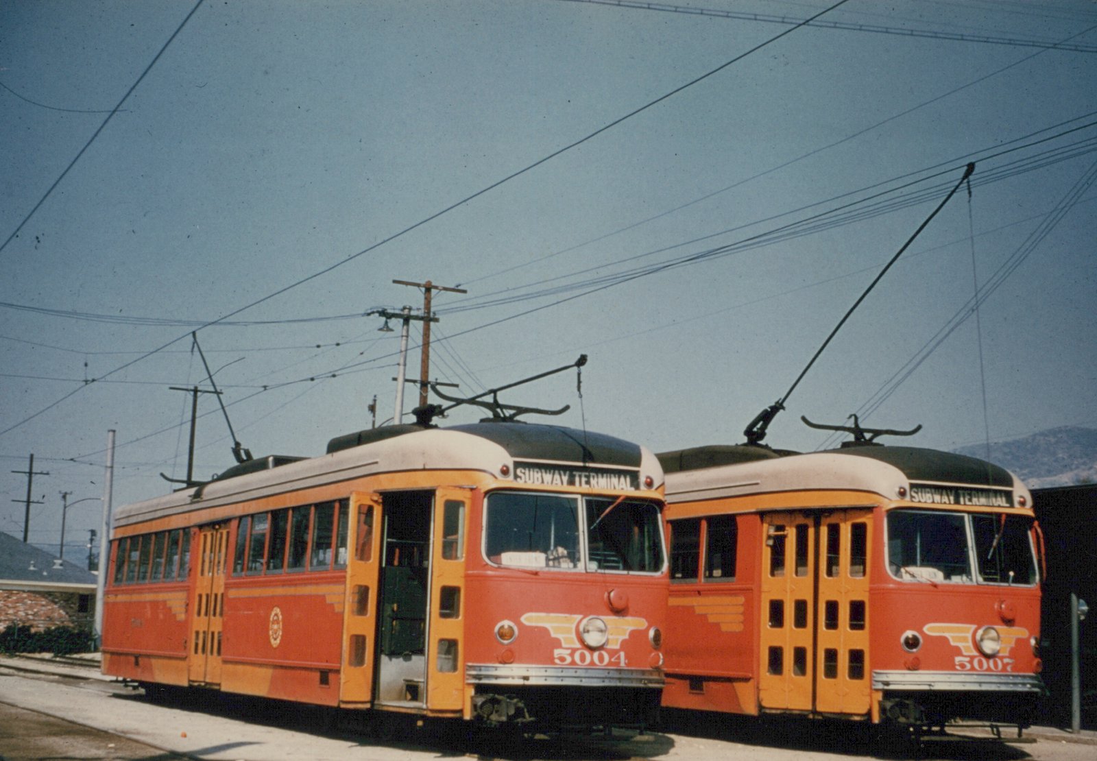 Two red and orange electric cars with yellow winged emblems on their fronts stand side by side under overhead wires, signed Subway Terminal, with hills in the background.