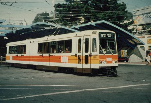 A white articulated light rail car with orange and yellow stripes rounds a curve on a paved plaza under a tangle of overhead wires, in front of a blue sawtooth canopy and tall trees.