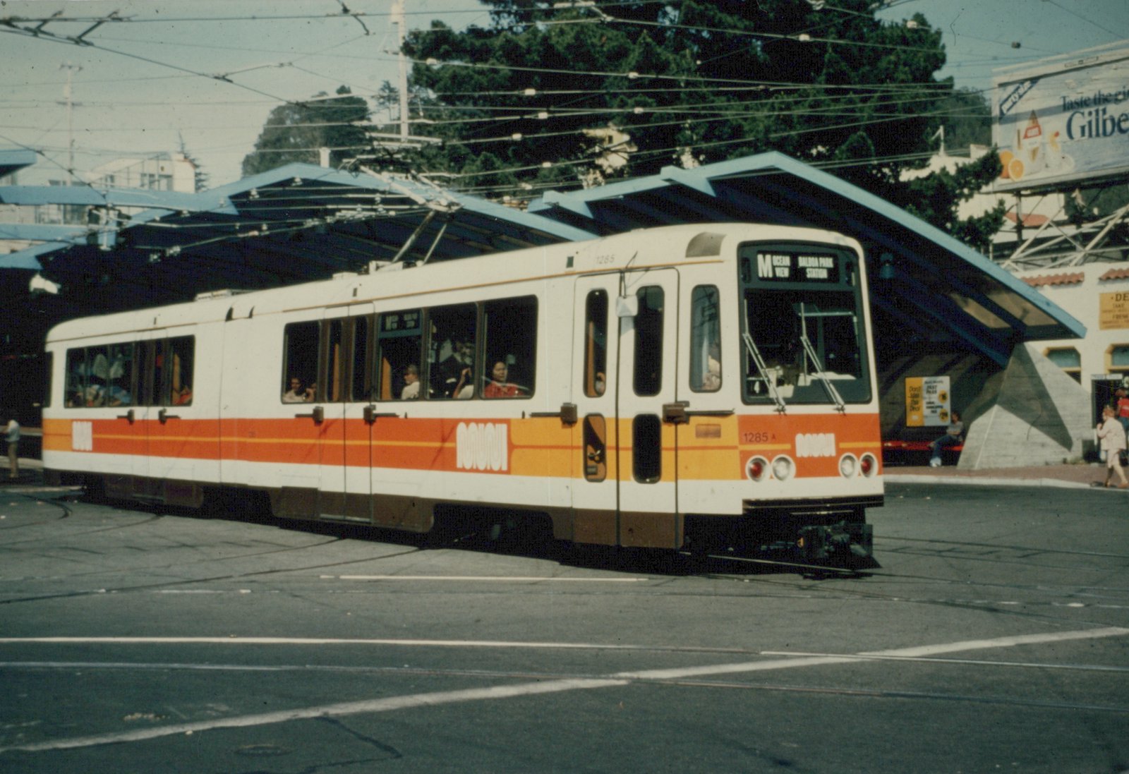 A white articulated light rail car with orange and yellow stripes rounds a curve on a paved plaza under a tangle of overhead wires, in front of a blue sawtooth canopy and tall trees.