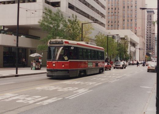 A red and white streetcar signed 504 Dundas West Station runs on street track past tall office buildings, with cars, taxis, pedestrians and a sidewalk food cart nearby.