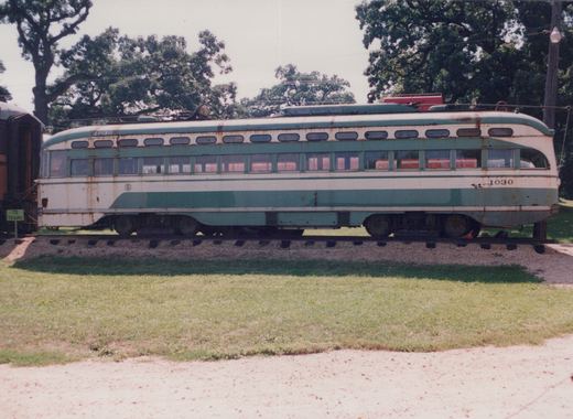 Side view of a streamlined cream and green streetcar numbered 1030, rusted and faded, standing on track in front of a lawn and large trees, with a TO TRAINS sign at the left.