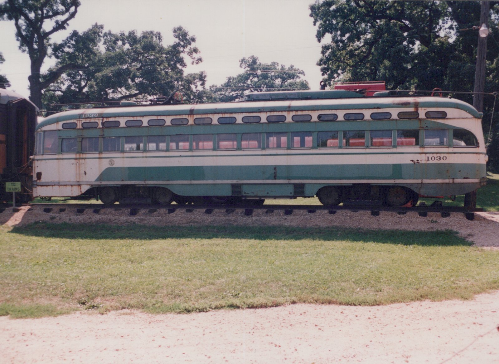 Side view of a streamlined cream and green streetcar numbered 1030, rusted and faded, standing on track in front of a lawn and large trees, with a TO TRAINS sign at the left.