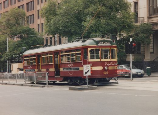 A maroon and gold tram with a single trolley pole and CITY CIRCLE in its roof sign, stopped on a tree-lined city street next to a safety island with a KEEP LEFT sign. Office buildings and a red traffic light are in the background.