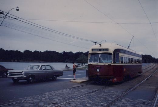 A maroon and cream streetcar numbered 4675 with the sign HUMBER QUEEN stops at a concrete island on paved double track, while a grey sedan passes on the road and a pond and trees are in the background under an overcast sky.