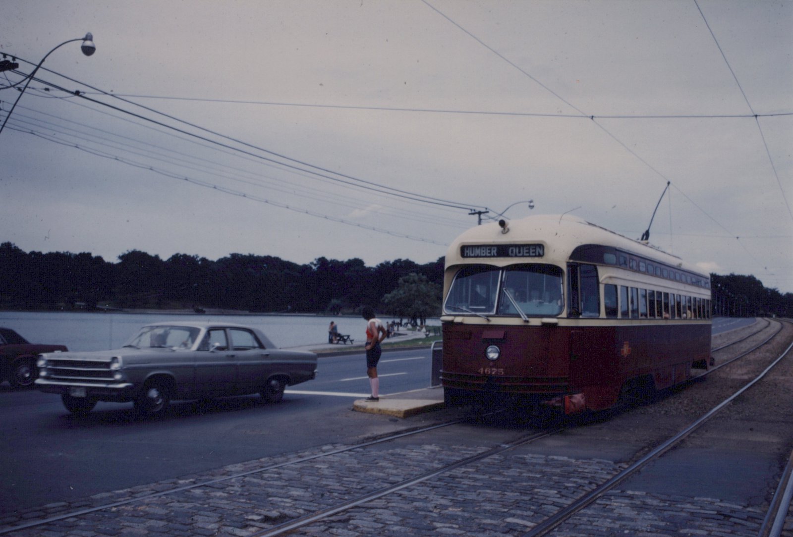 A maroon and cream streetcar numbered 4675 with the sign HUMBER QUEEN stops at a concrete island on paved double track, while a grey sedan passes on the road and a pond and trees are in the background under an overcast sky.