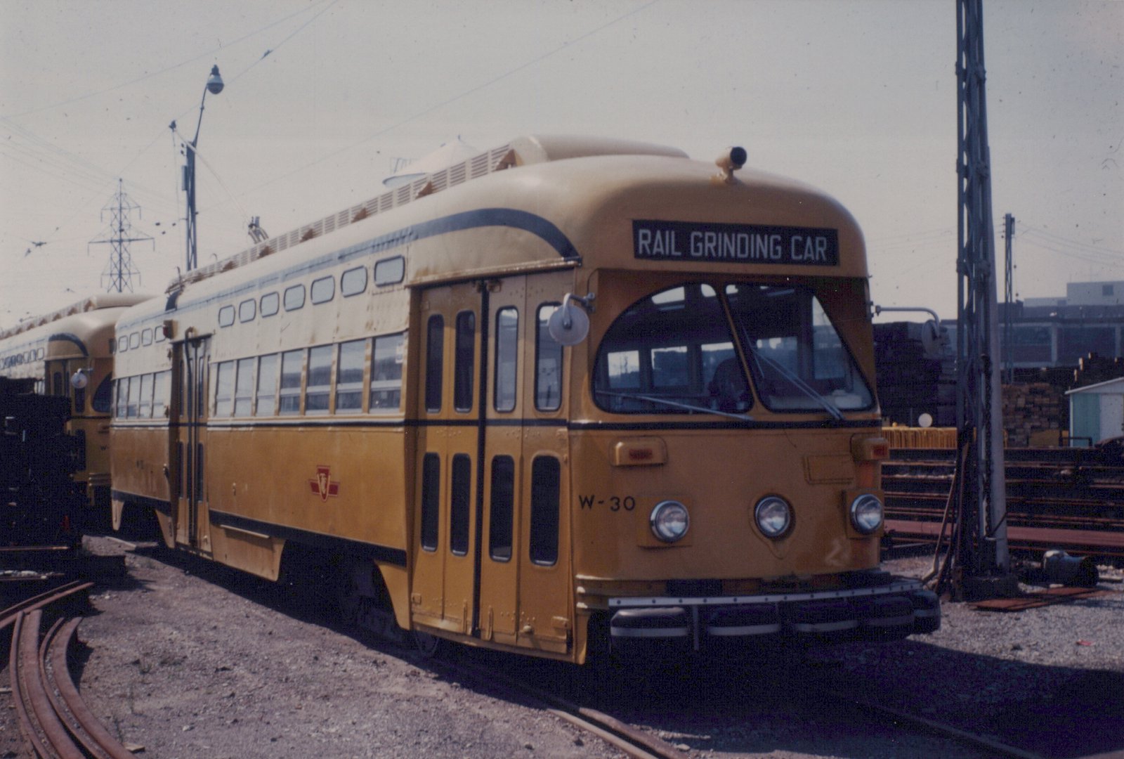 A yellow streamlined streetcar with RAIL GRINDING CAR on its destination sign and W-30 on its front stands on yard track, with a second yellow streetcar behind it and stacks of rail and ties to the right.