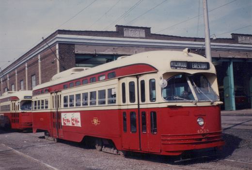 A red and cream streetcar numbered 4558 stands outside a brick carhouse with open bays, with a second streetcar behind it on the left under overhead wires.