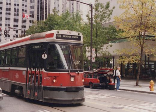 A red and white streetcar numbered 4120 with a sign reading 504 Broadview Station travels a downtown street past a parked red station wagon, a man standing in the road and trees with yellowing leaves.