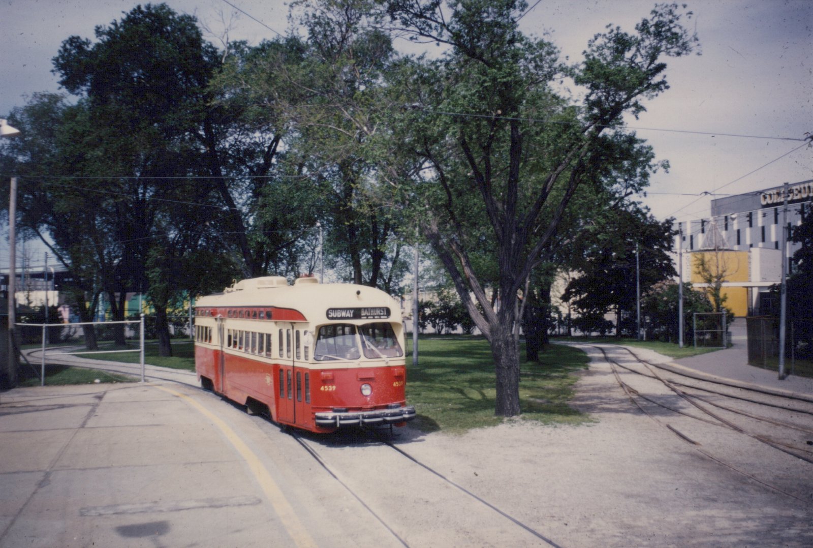 A red and cream streetcar with a destination sign reading SUBWAY BATHURST rounds a curved loop track among tall trees, with a large building lettered COLISEU at the right.