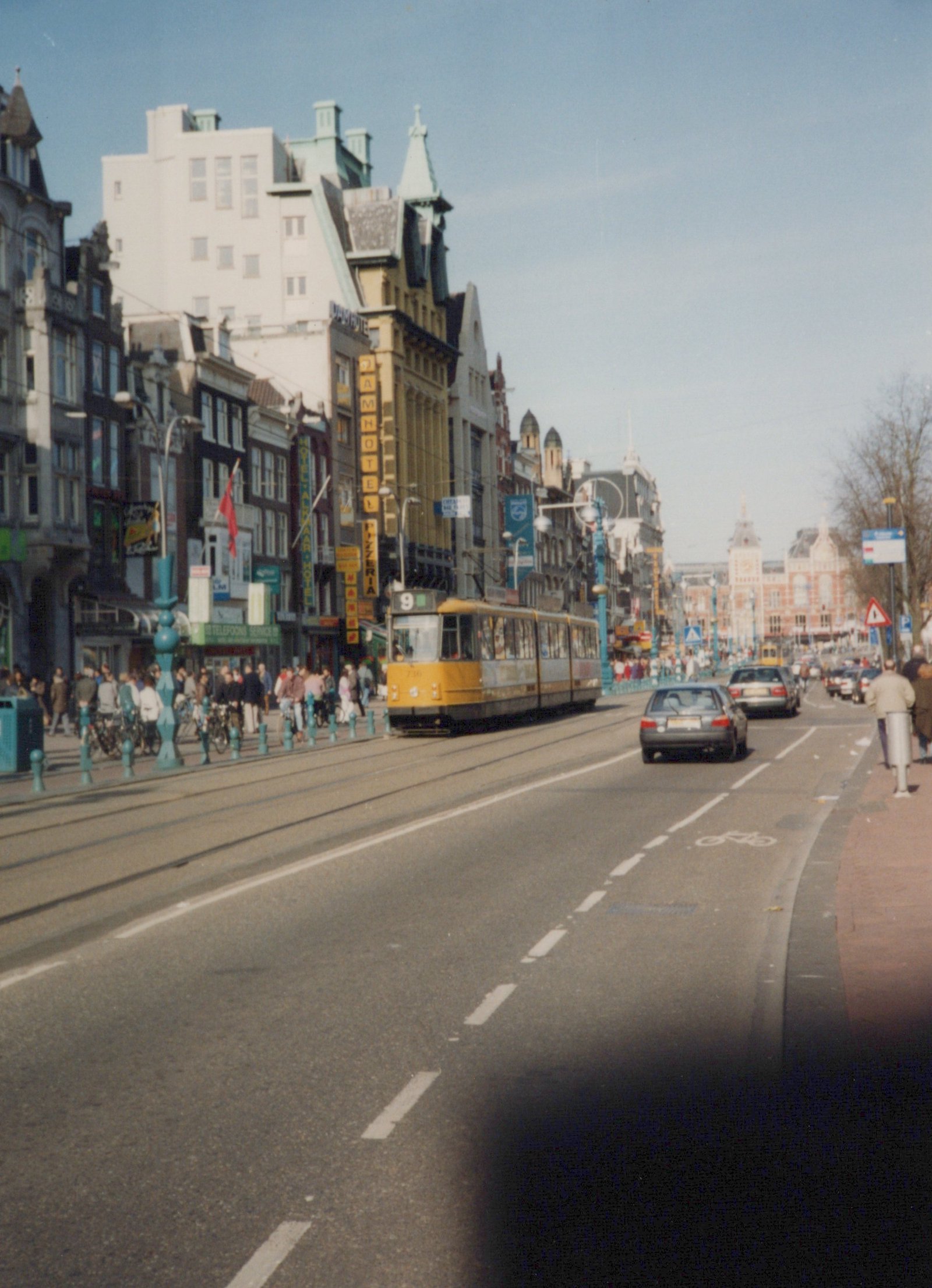 A yellow articulated tram showing route 9 travels along tracks in a wide city street lined with tall old buildings and hotel signs, as cars drive in the adjacent lane and crowds walk on the sidewalk.